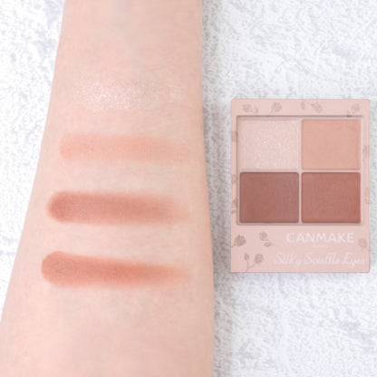 Swatches of Canmake Silky Flare Eyes matte type palette on an arm, displaying gold, bronze, and copper shades alongside the palette.