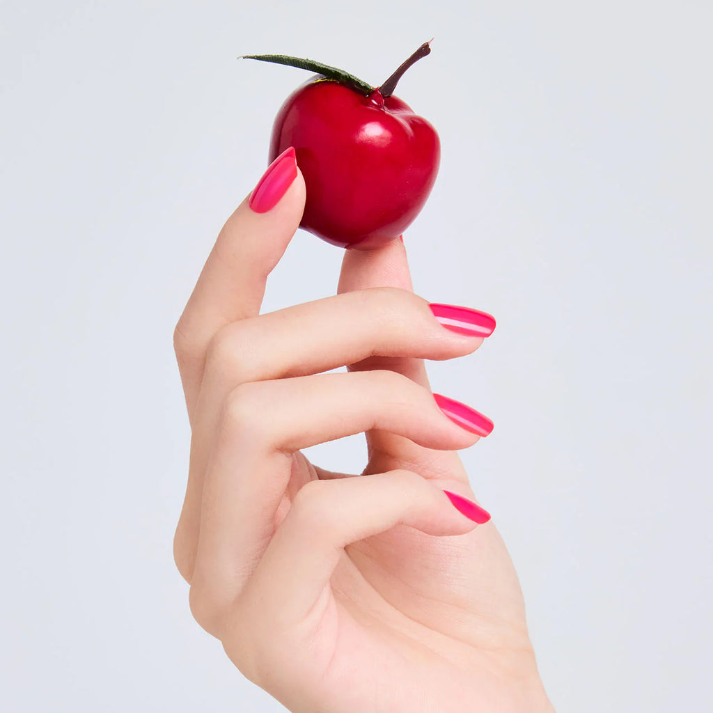 Hand with nails painted in vibrant pink Elégance Lacquer, holding a red cherry, showcasing a glossy and playful finish.